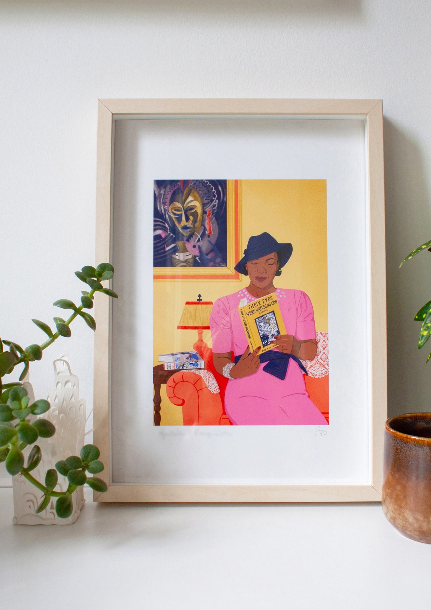 Framed illustrated portrait of Zora Neale Hurston in her living room, reading her book. Behind here is a painting by Lois Mailou Jones, another important Black woman of the Harlem Renaissance.